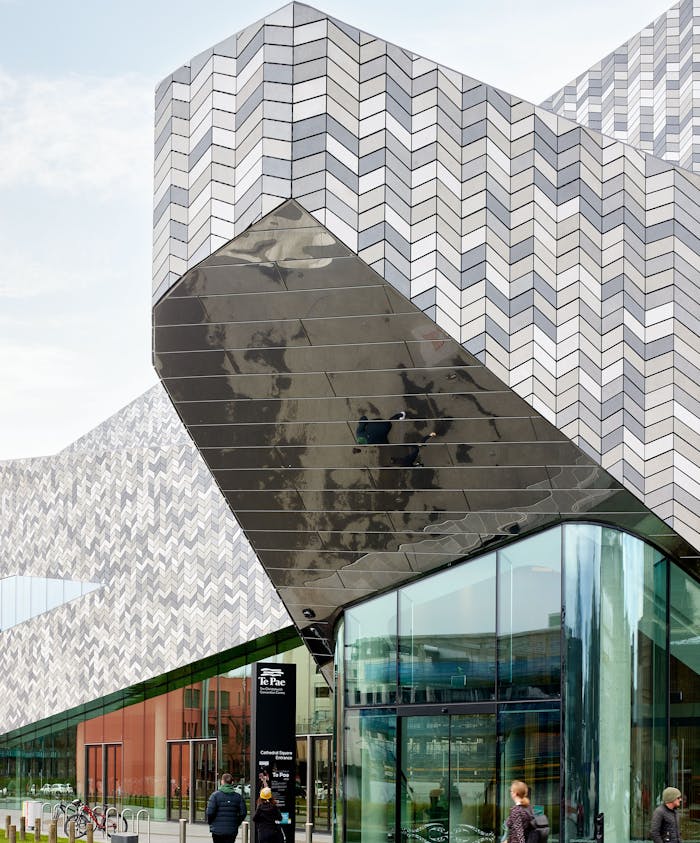 A striking modern building features a mix of angled, patterned surfaces in gray, black, and reflective materials. People walk by as they approach the entrance marked “Te Pae.”